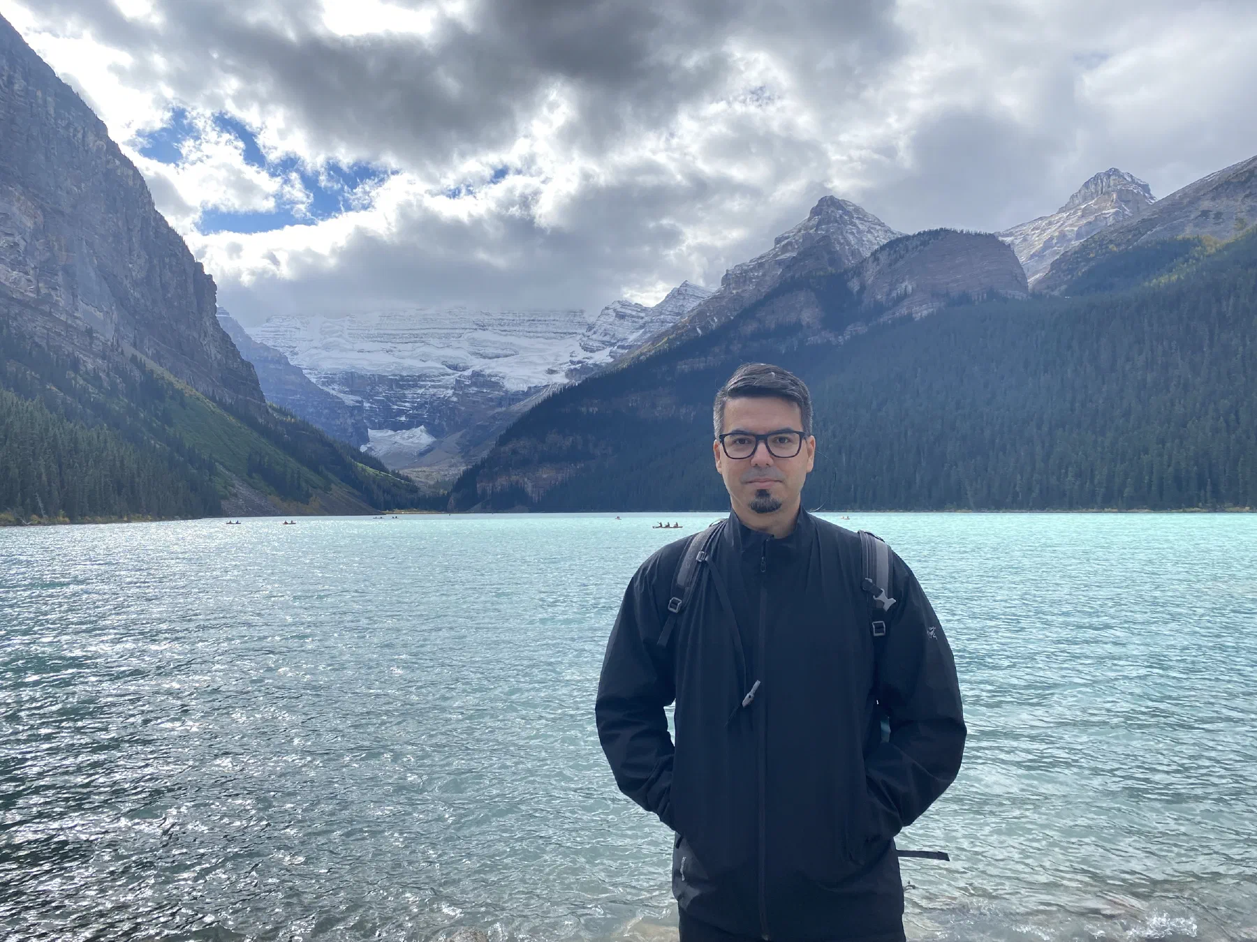 Victor standing by the turquoise water of Lake Louise, with forested mountains and a glacier behind him
